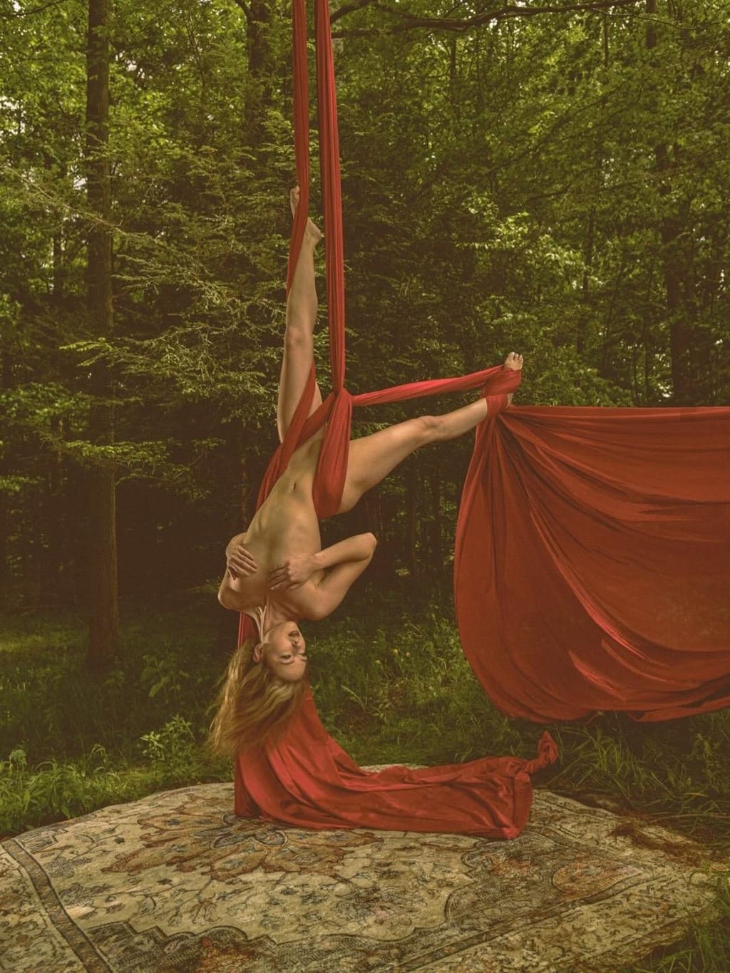 Lana West performing an aerial silks split pose wrapped in red fabric in a forest.
