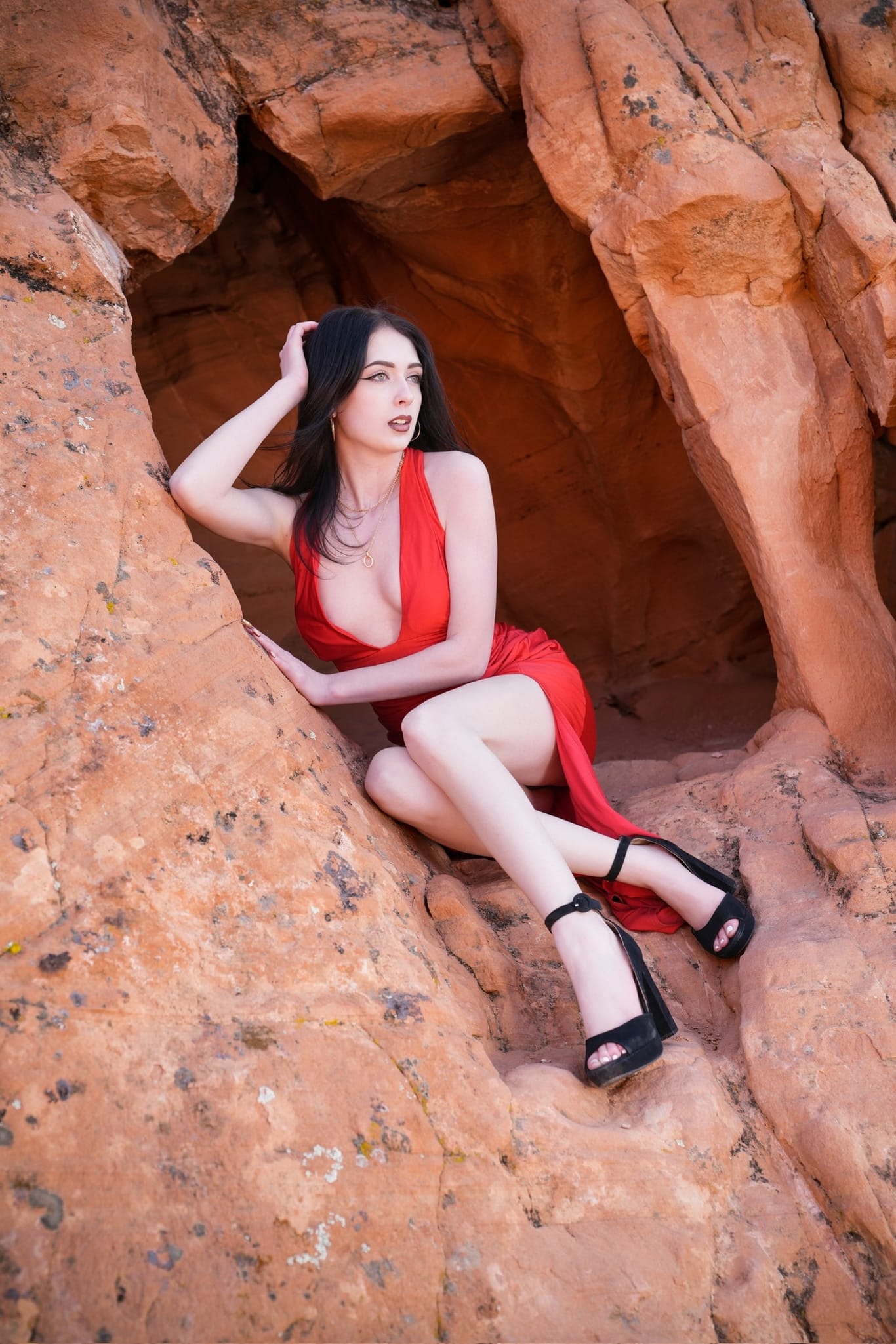 Lana West in a plunging red dress seated in a red-rock desert cave.